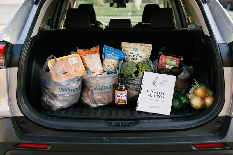 Walmart bags of chicken, rice, cauliflower rice, broccoli, ground beef, and avocados in the back of an SUV, with the cookbook