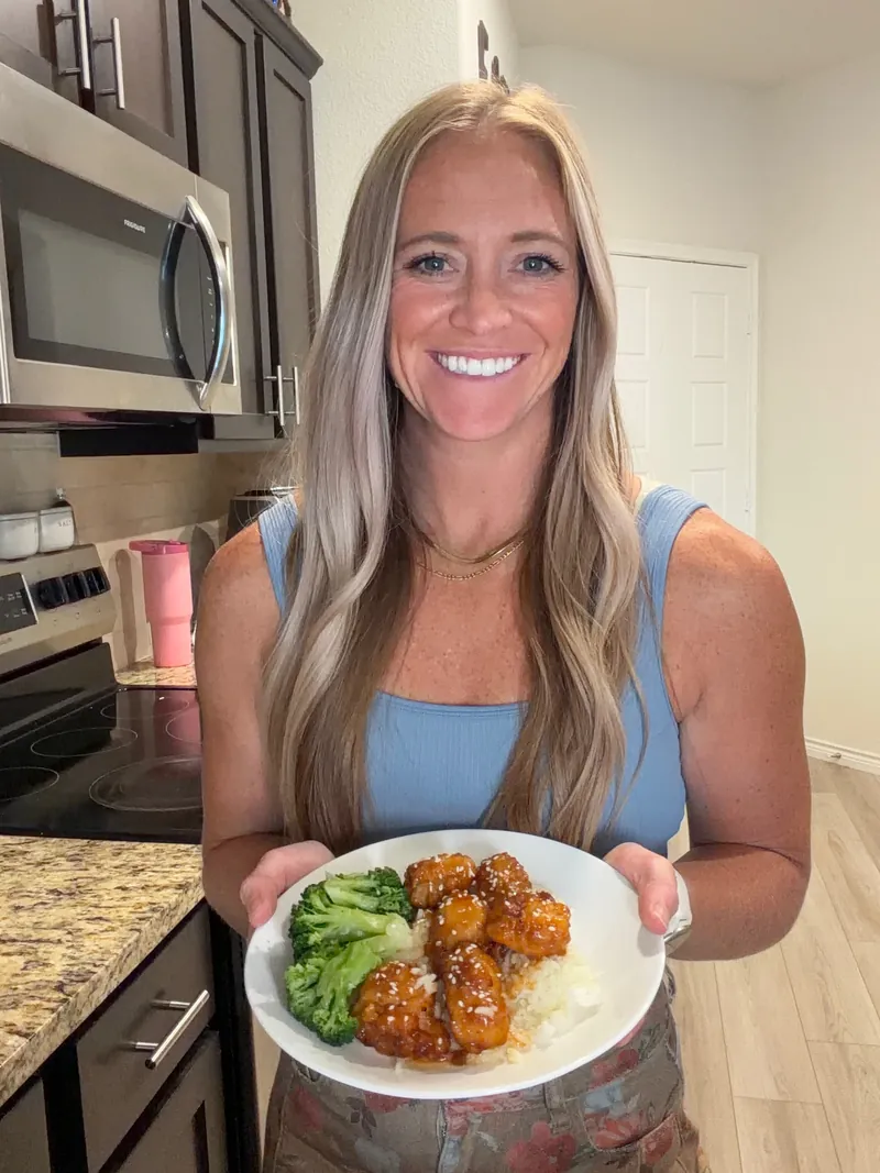Miranda in her kitchen, holding a plate of orange chicken, rice, and broccoli